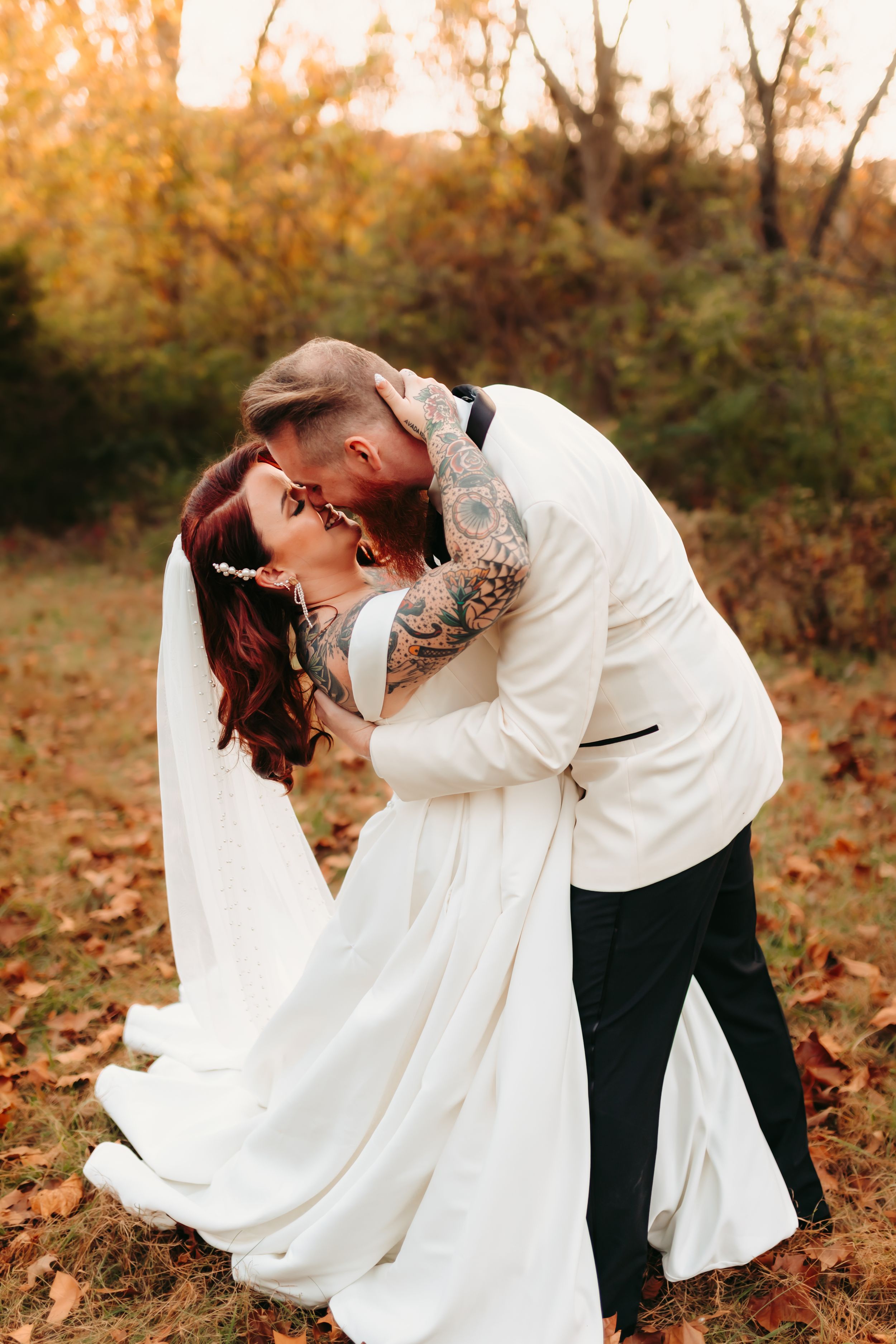 Romantic couple in wedding attire share intimate kisses in a golden autumn forest setting with fallen leaves.