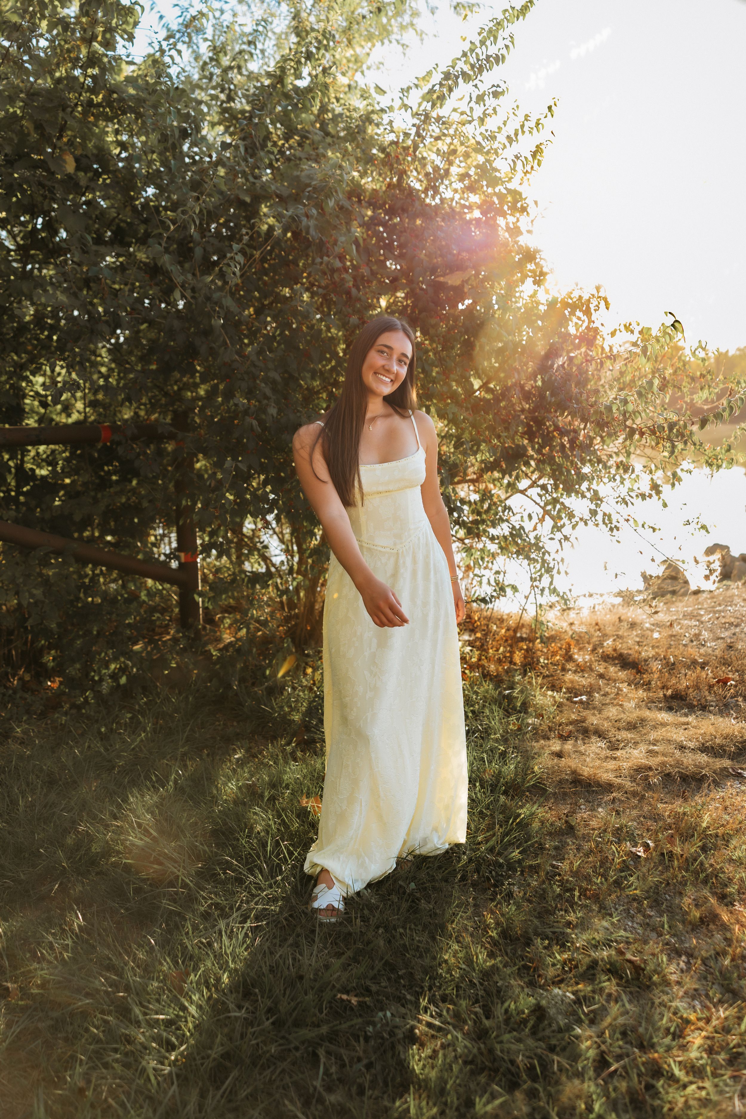 A person in a flowing white sundress stands in a sunlit field with trees during golden hour.