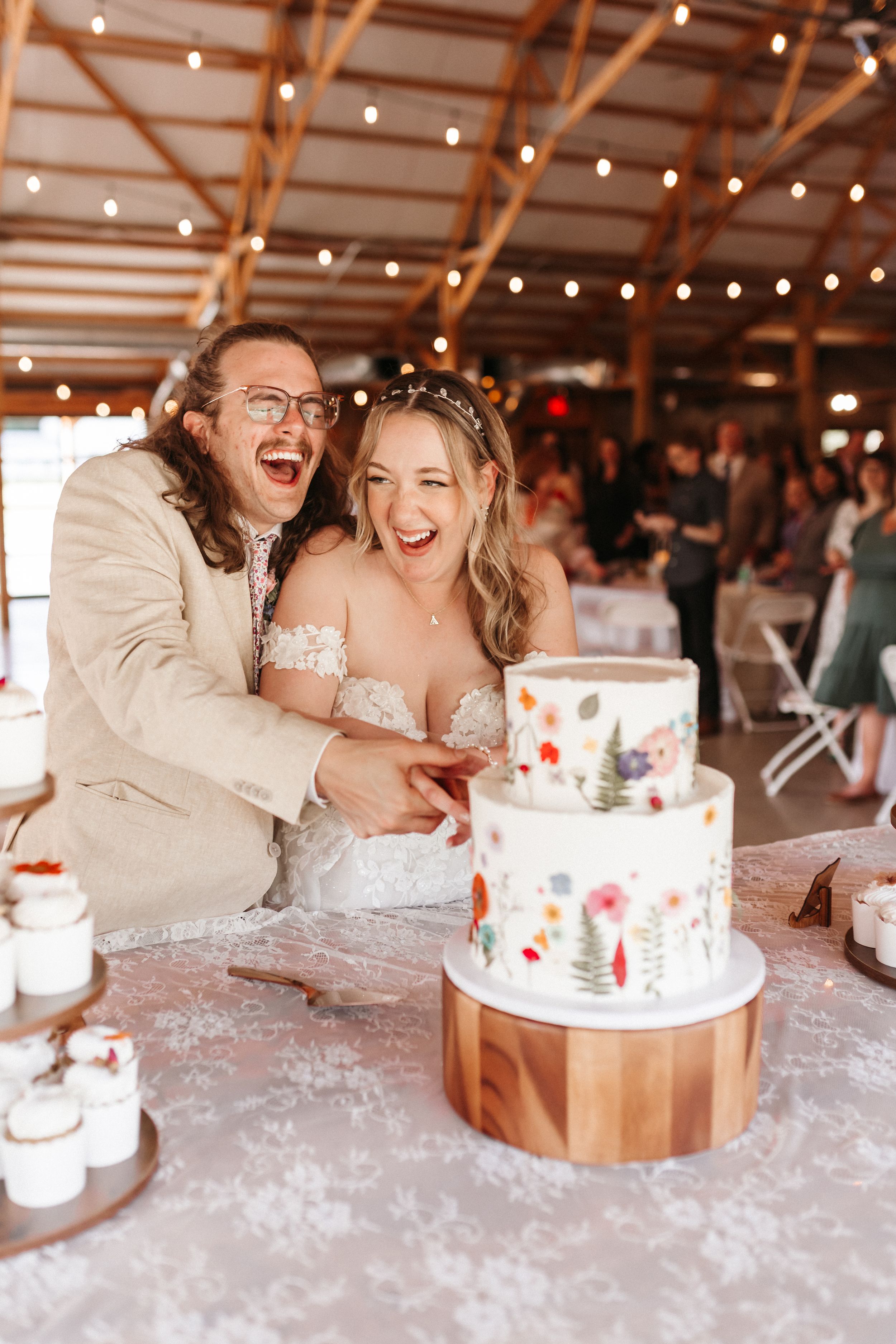 A couple laughs joyfully together while sitting at their wedding reception table with decorated cakes.