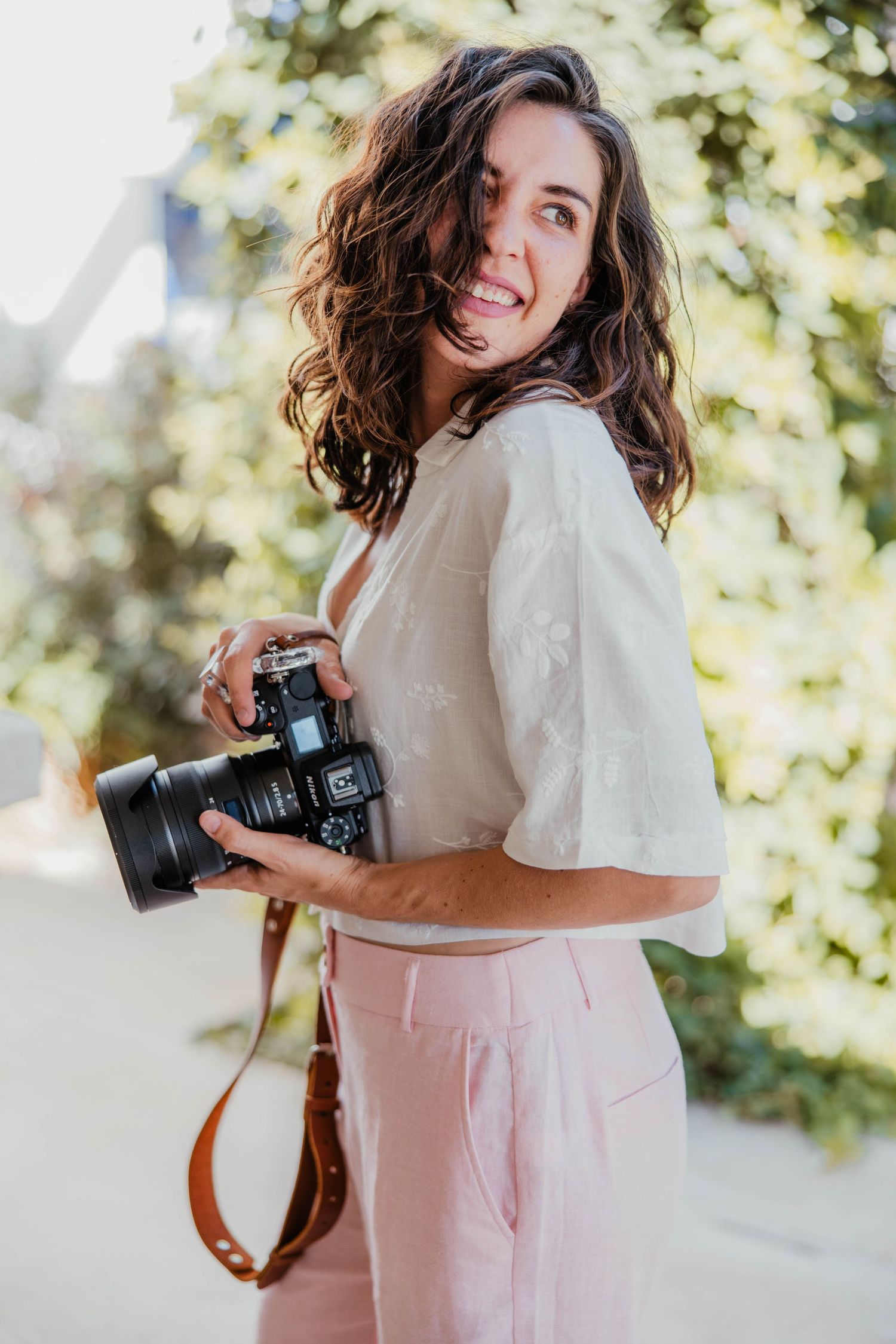 Photographer in casual attire smiling while holding professional camera outdoors.