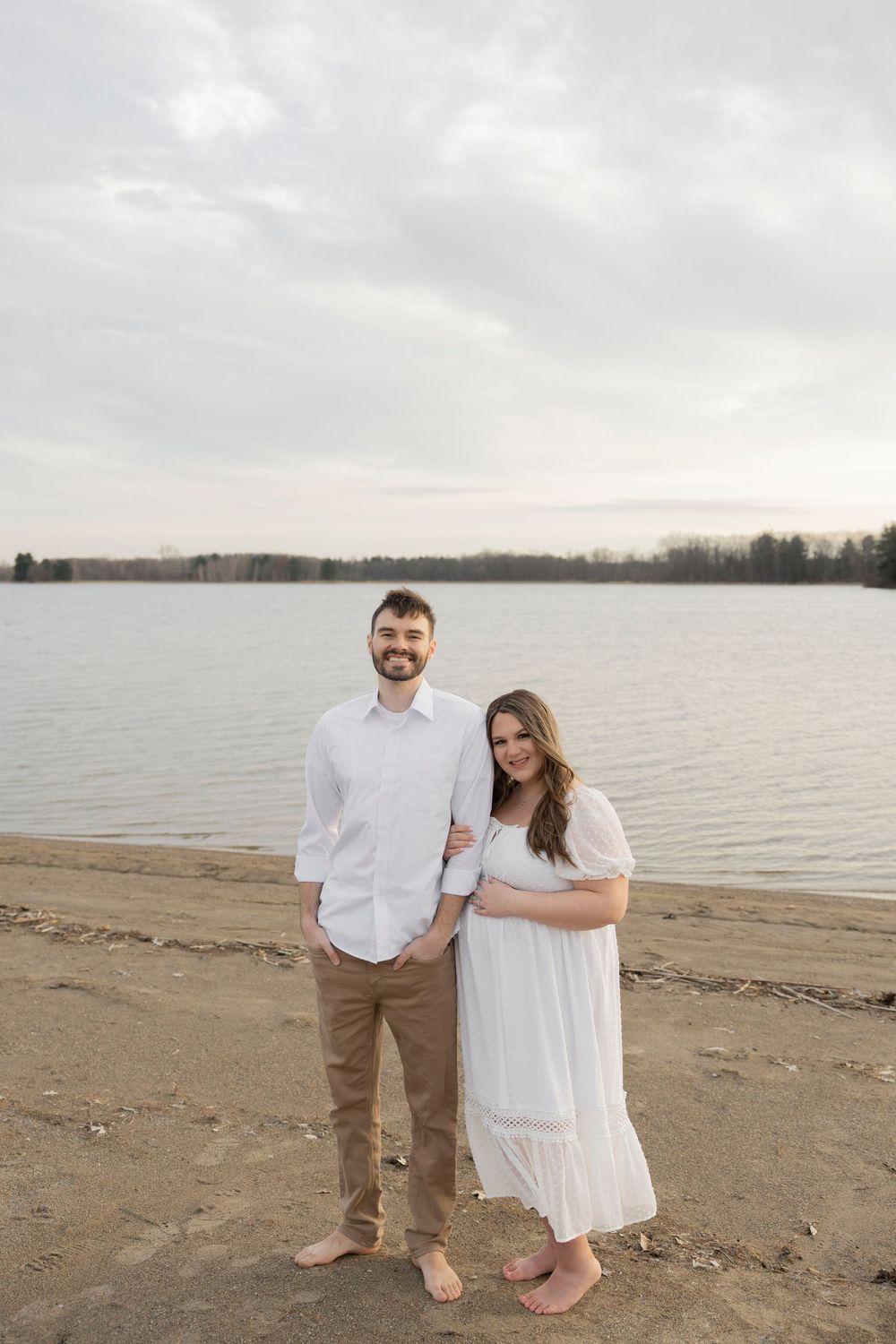 Pregnant couple in white outfits standing barefoot on sandy beach by calm lake, posing for romantic outdoor portrait.
