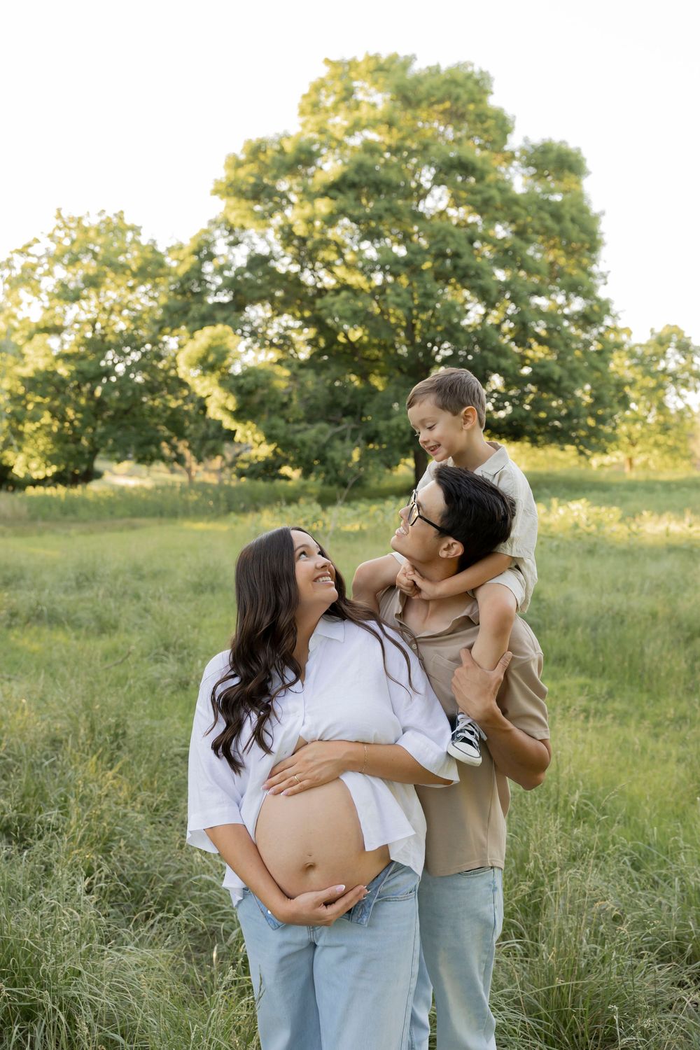 family smiling as they play in a feild in lake Orion Michigan while toddler boy is only dads shoulders and mom holds her pregnant belly