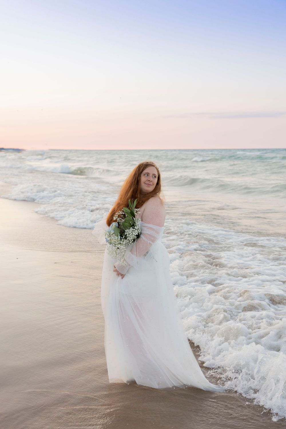pregnant woman in white tulle gown holds bouquet at Lake Huron shore during pastel sunset in michigan
