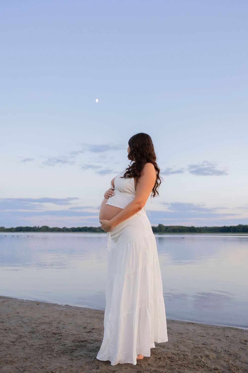 Pregnant woman in white dress stands on sandy beach at dusk, cradling her belly while gazing at calm water atop stoney creek metropark