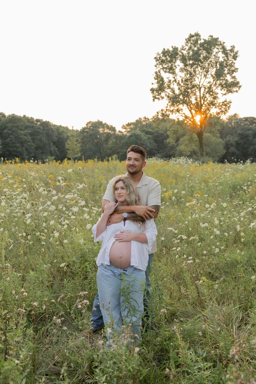 Couple embracing in a wildflower field at sunset, pregnant woman showing baby bump in soft golden light.