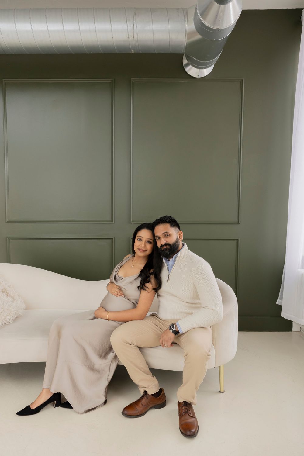 A pregnant couple in neutral outfits poses together on a white couch against a dark green wall at LMP collective in Pontiac michigan