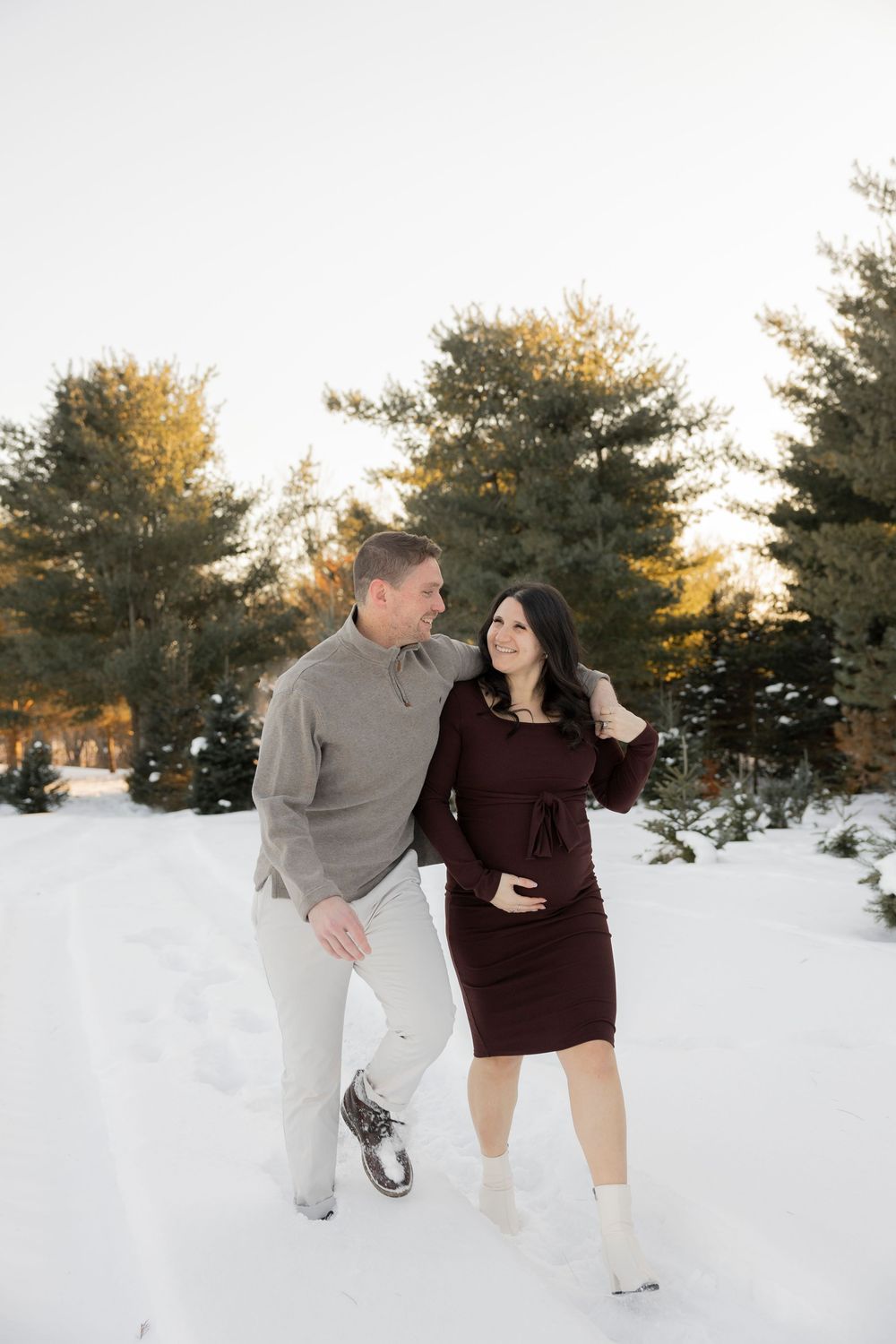 A pregnant couple in winter attire poses together in a snowy landscape with evergreen trees and golden sunlight in lake Orion michigan