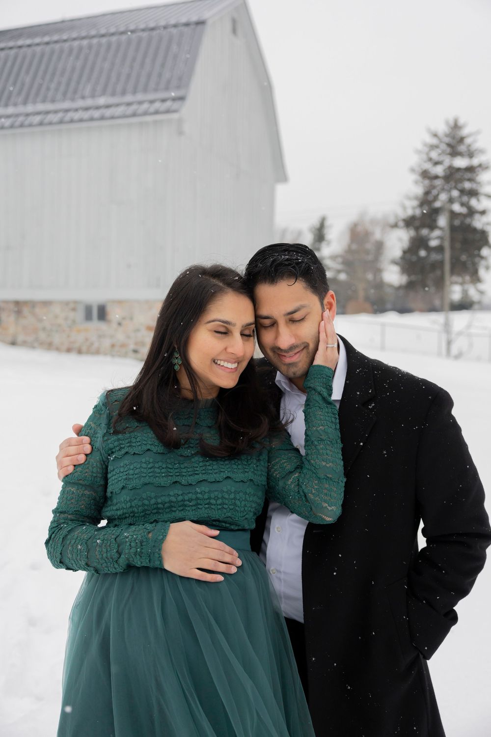 A pregnant couple embraces in a snowy winter setting in front of a barn at troy farm park near Rochester hills michigan for a photoshoot