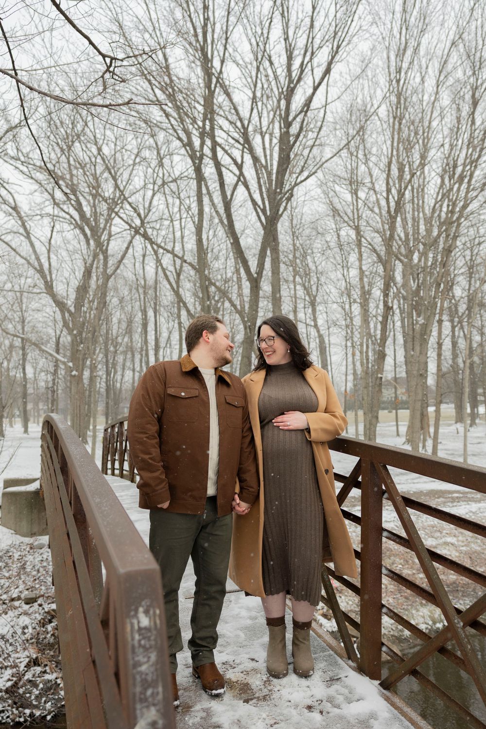 A pregnant couple stands together on a snowy wooden bridge in troy michigan in a winter forest setting for winter maternity photos