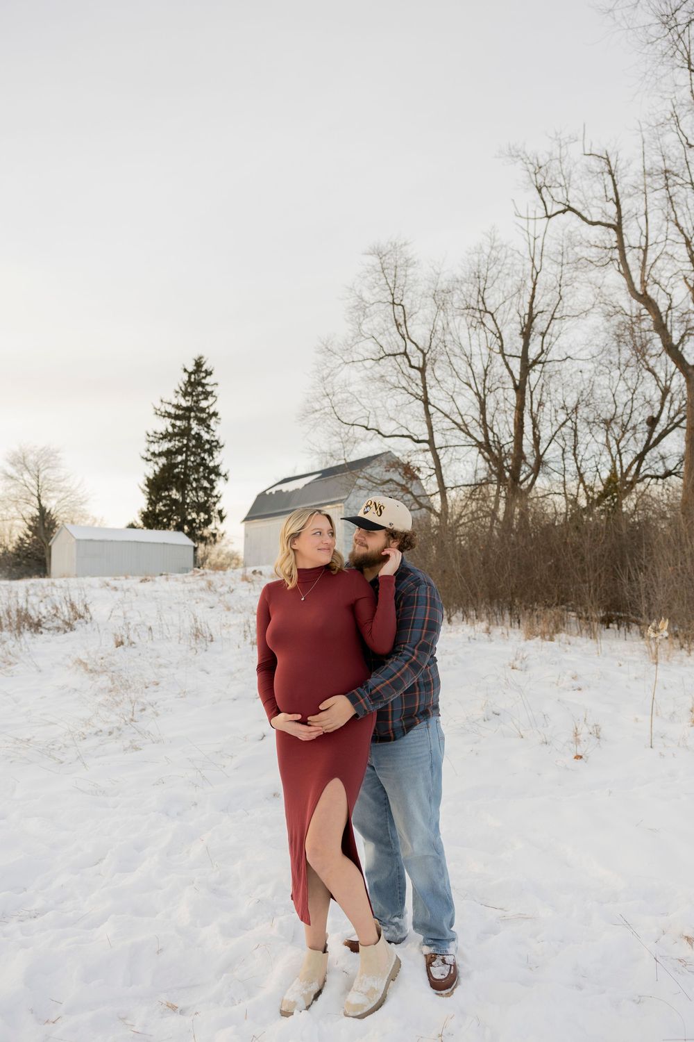 A pregnant couple embraces in a snowy winter landscape with a white barn creating a romantic outdoor scene in troy Michigan near Rochester