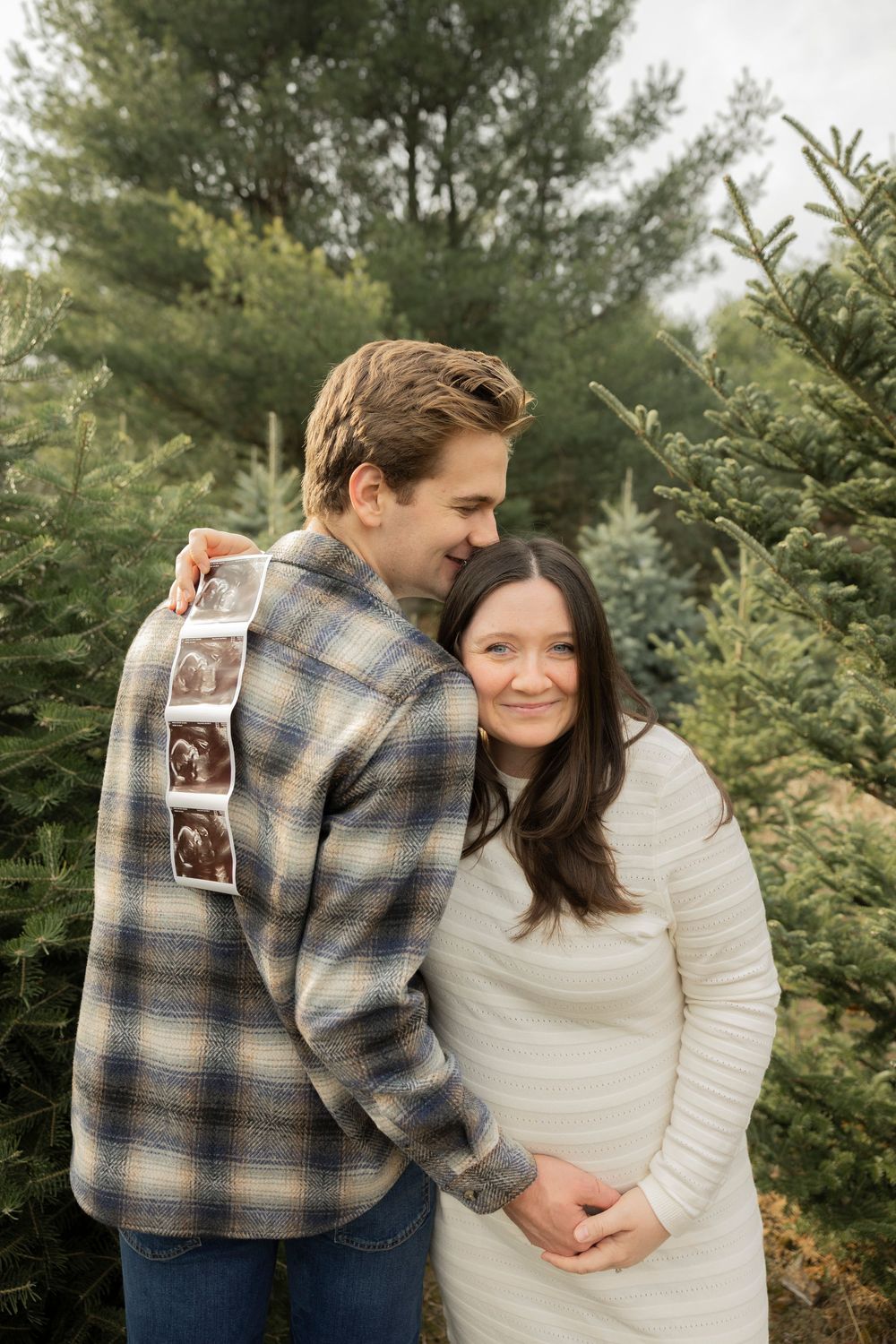A pregnant couple shares tender moments among evergreen trees while holding an ultrasound photo during a pregnancy announcement shoot.