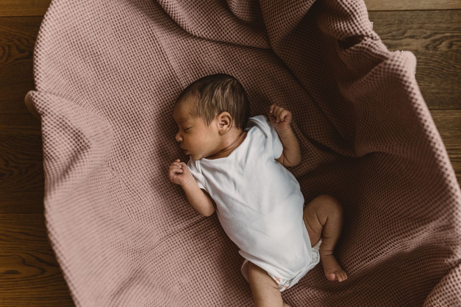Newborn baby in white onesie lying in wicker bassinet with sage green waffle blanket.