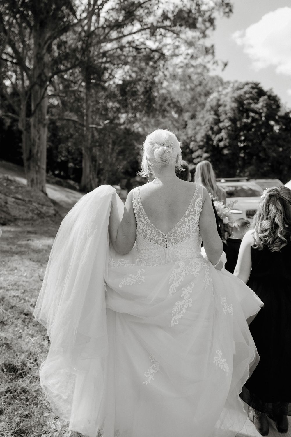 Black and white photo of a bride in a lace wedding gown and veil walking with bridesmaids outdoors.