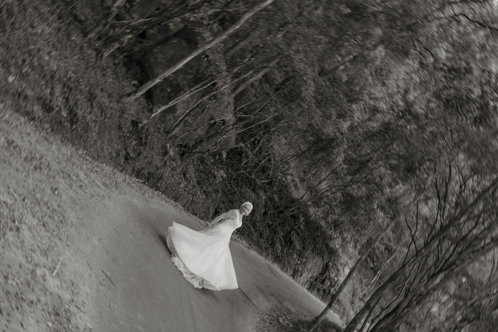 Black and white aerial photo of a woman in a white dress walking along a curved path surrounded by trees.