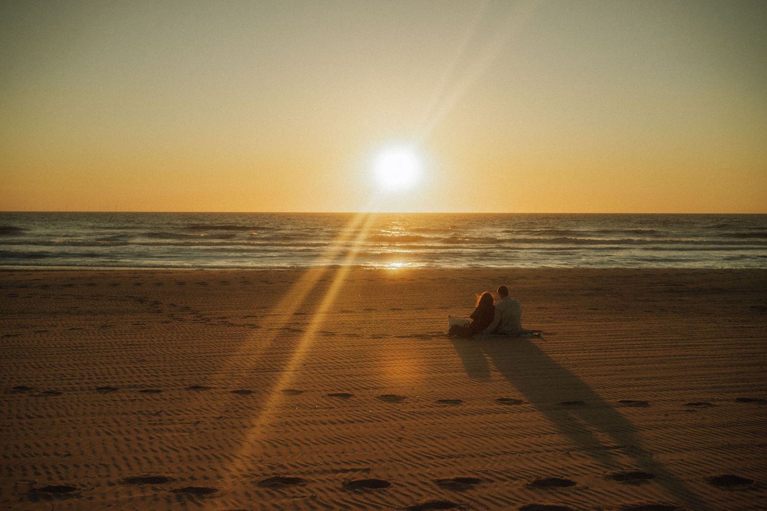 Two people sit together on a sandy beach at sunset, casting long shadows as golden sunlight glows over the calm ocean horizon. Netherlands