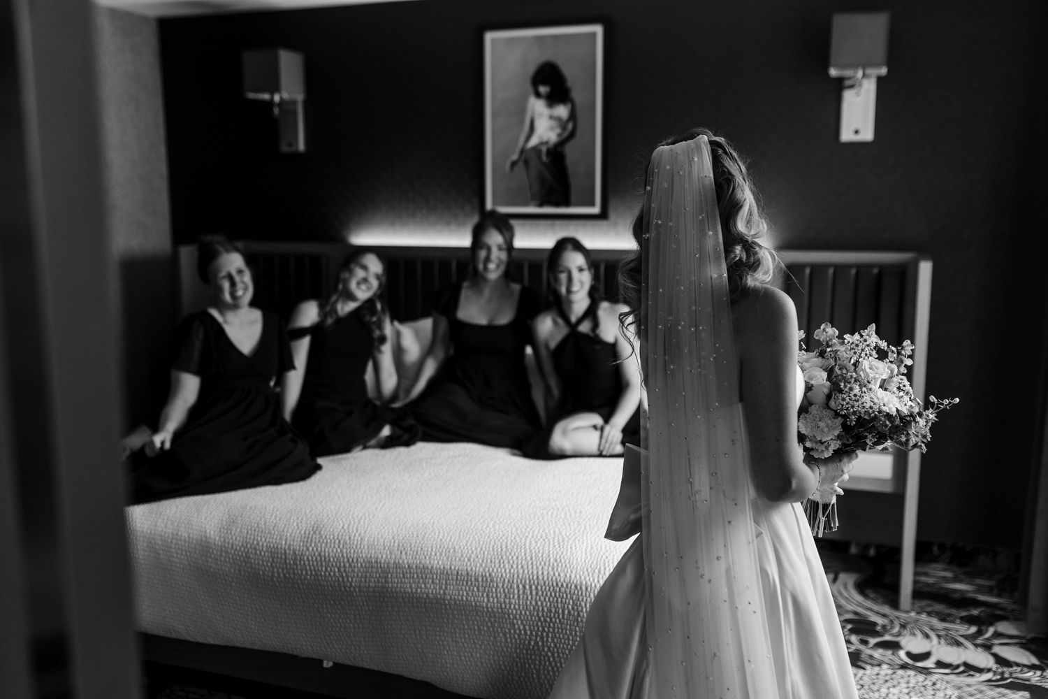 Bride in white gown shows bridesmaids her wedding dress in black and white hotel room photo.