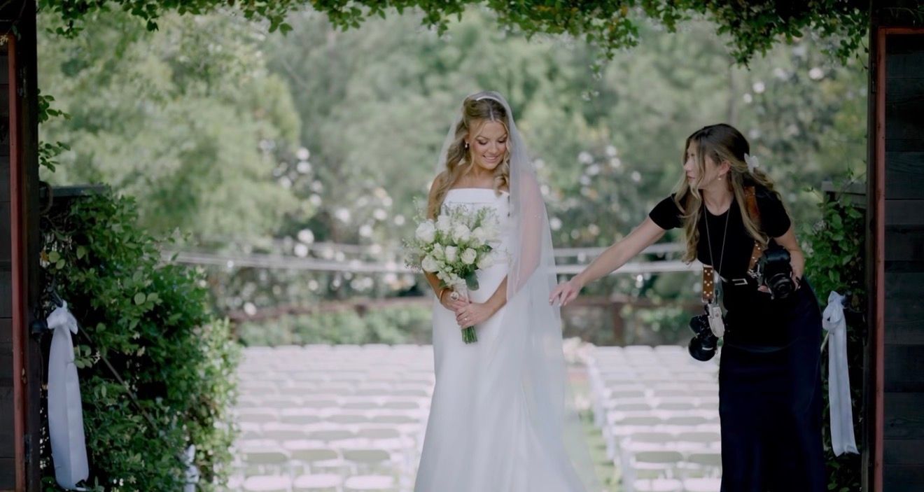 Elegant bride in white gown holding bouquet standing under a garden arch with a photographer nearby.