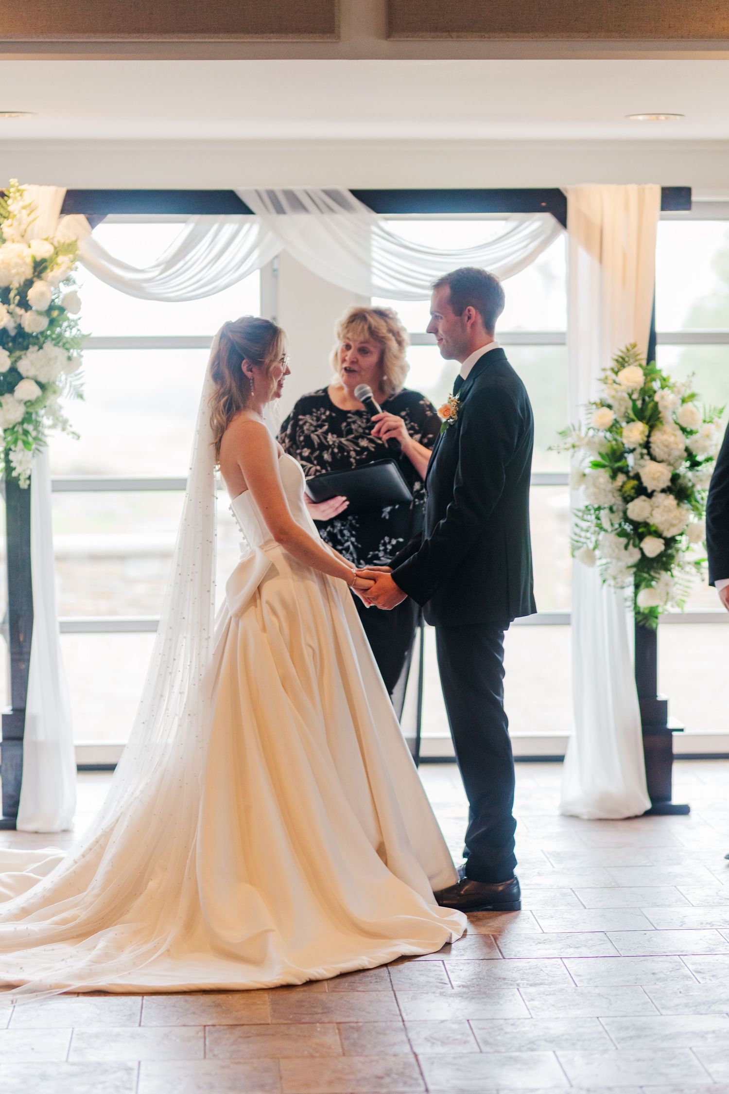 Bride and groom exchange vows at indoor wedding ceremony with white floral arrangements and draped backdrop.