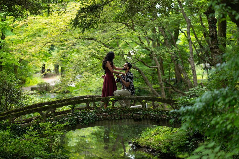 Man kneeling on one knee proposing to a woman in a red dress on a rustic wooden arched bridge in a lush green Japanese garden.