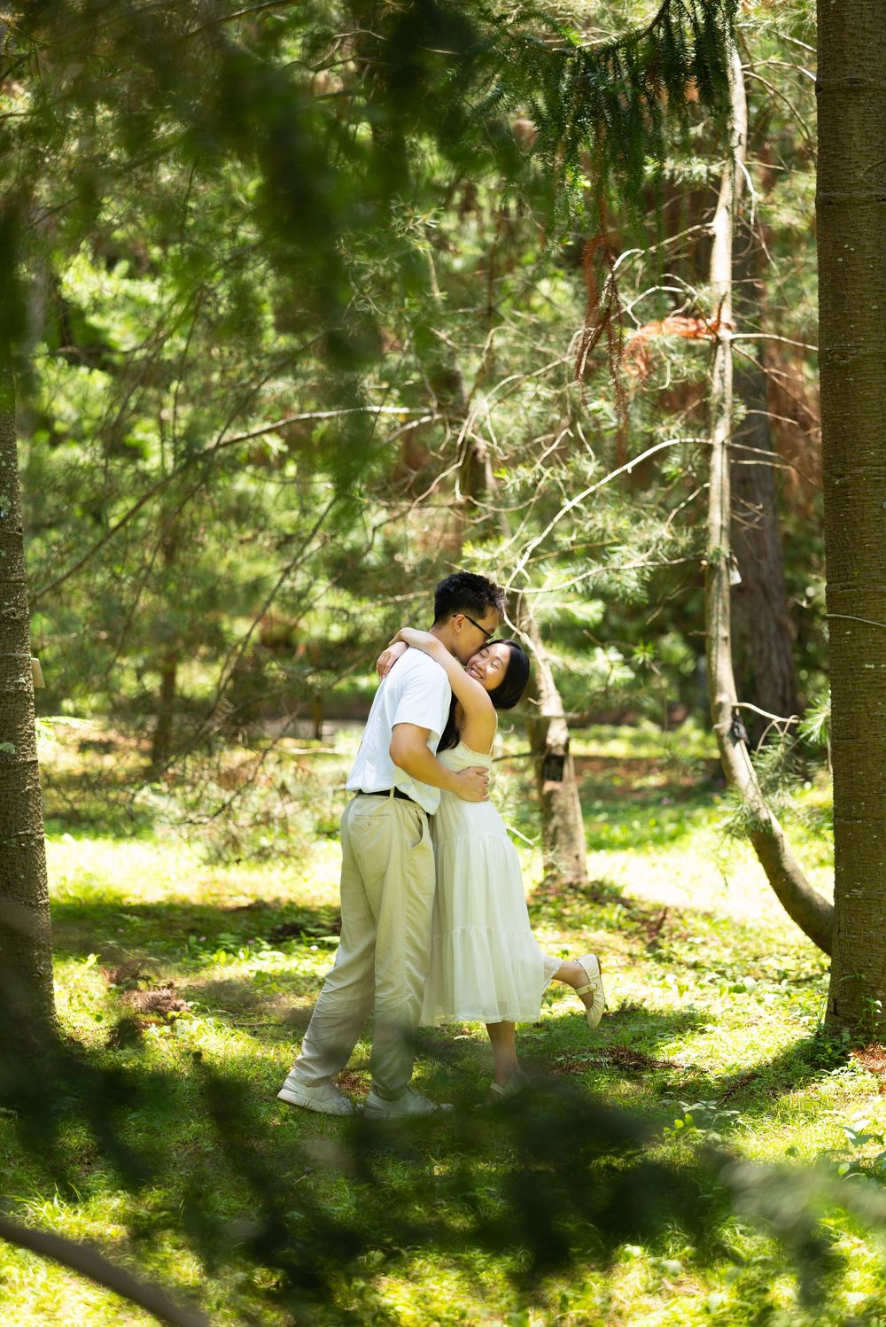 Couple in a loving embrace among tall pine trees in a sunlit forest, the woman in a white dress and the man in a white shirt.
