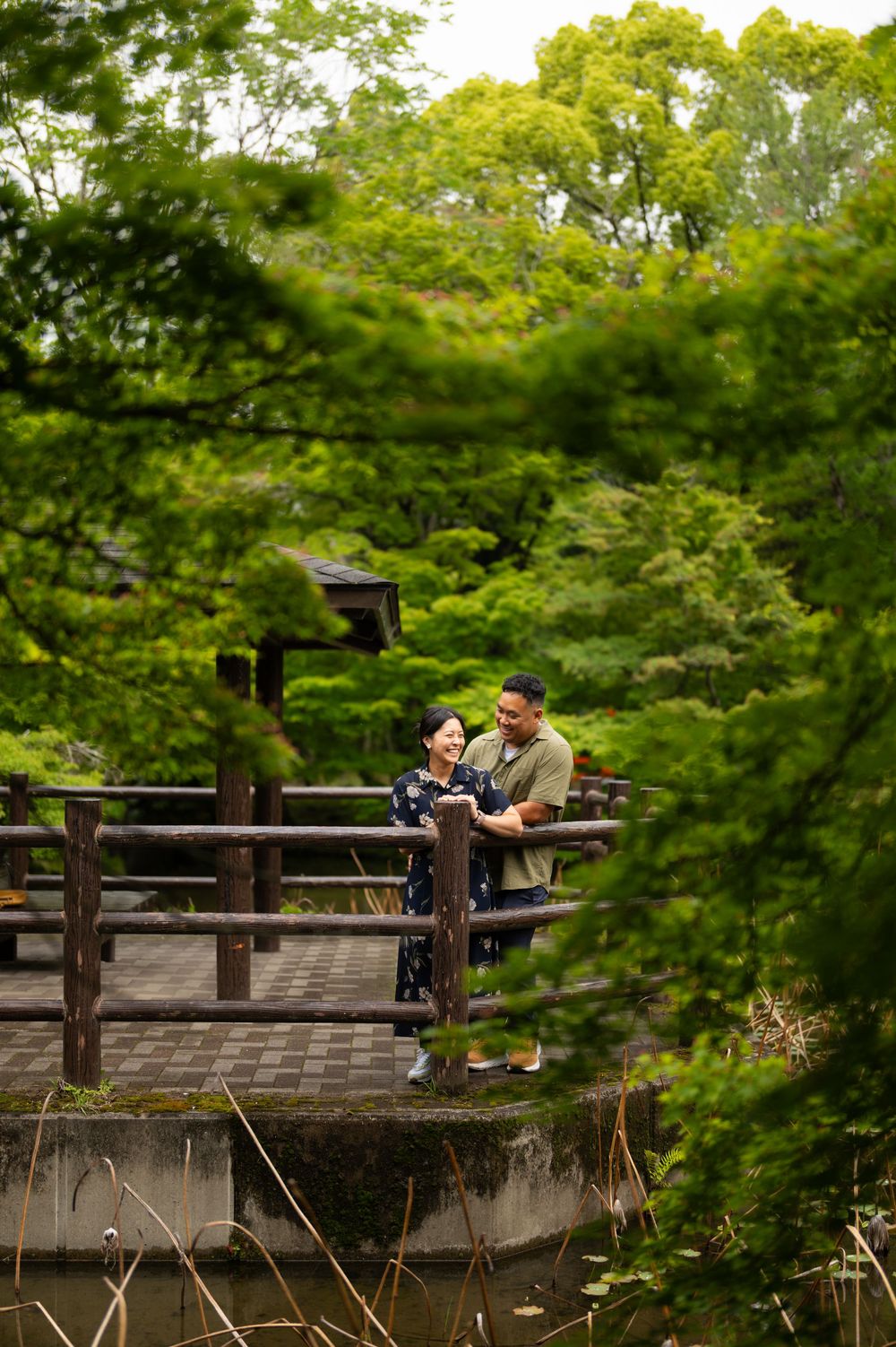 A smiling couple embraces on a wooden bridge in a lush Japanese garden, surrounded by vibrant green trees and a traditional pavilion.