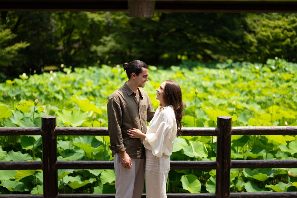 Romantic couple standing face to face beside a wooden railing, surrounded by lush green lotus leaves in a serene garden pond.