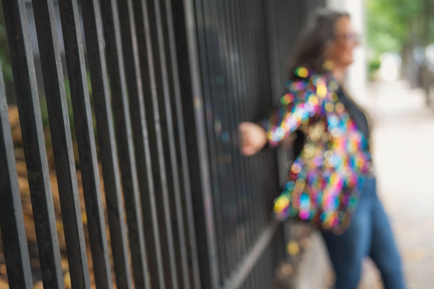 Blurred woman in a colorful sequin jacket and jeans stands beside black iron fence bars on a tree-lined city sidewalk.