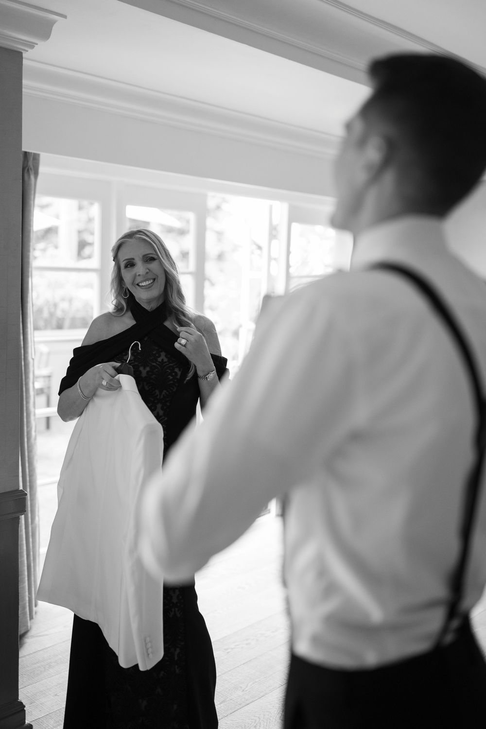 Black and white photo of a smiling woman holding a suit jacket for a man in suspenders during wedding preparations.
