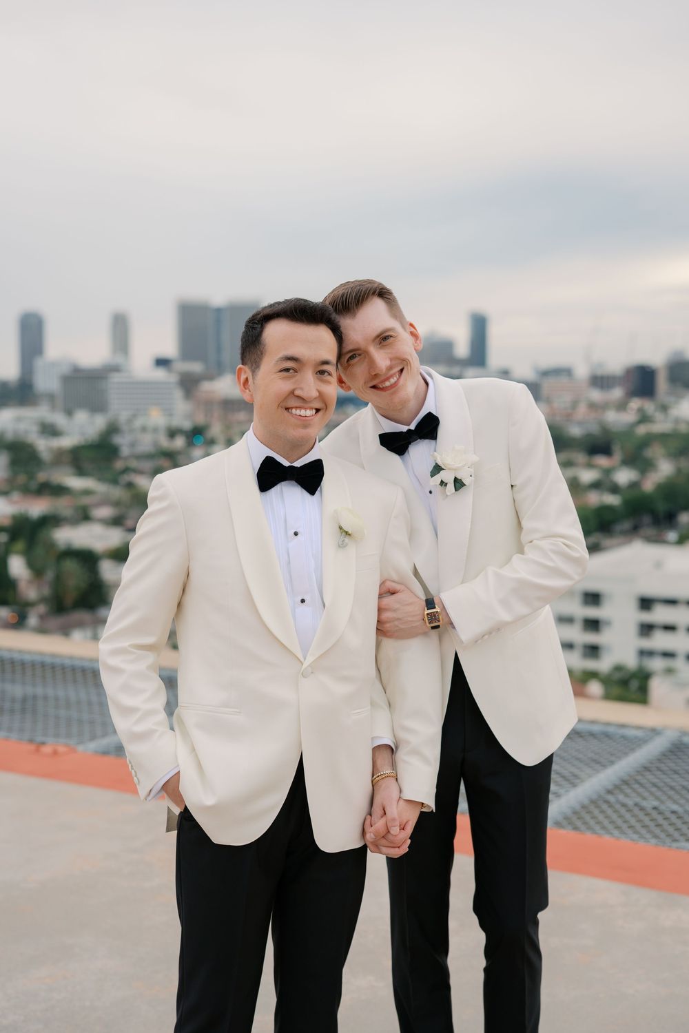 Two grooms in white tuxedo jackets and black bow ties pose together on a rooftop with a city skyline backdrop.
