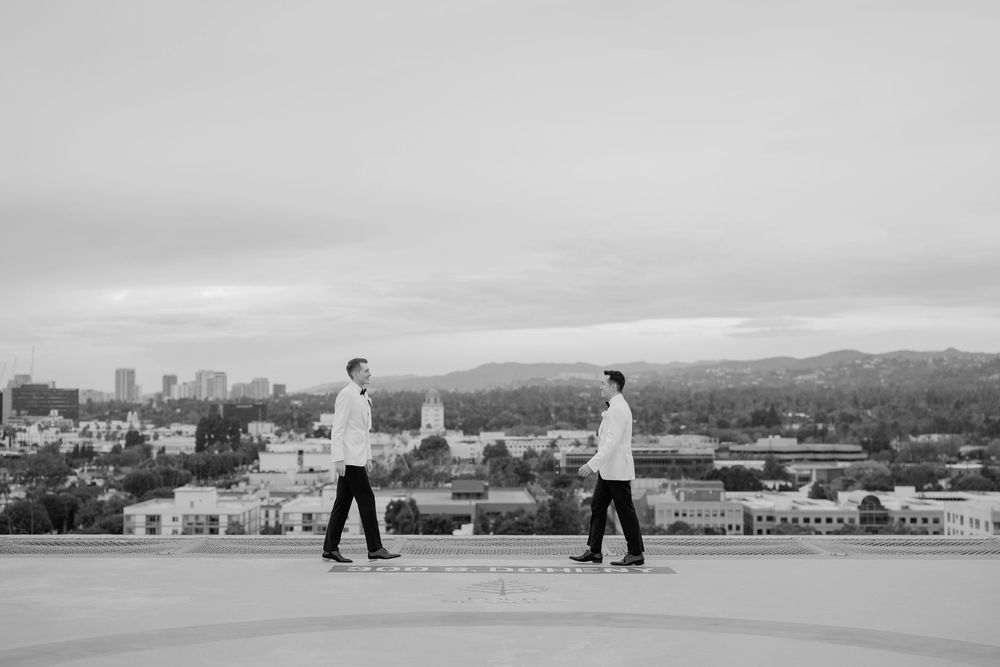 Two grooms in white shirts walk on a rooftop with a city skyline and mountains in the background.