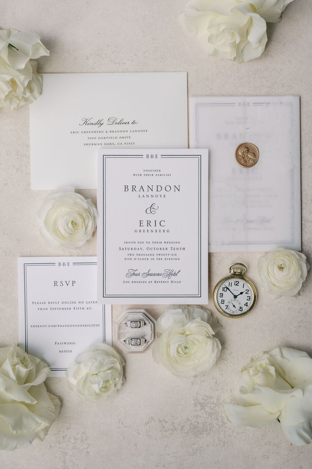 Elegant wedding invitation suite with white ranunculus flowers, gold wax seal, vintage pocket watch, and diamond ring on ivory background.