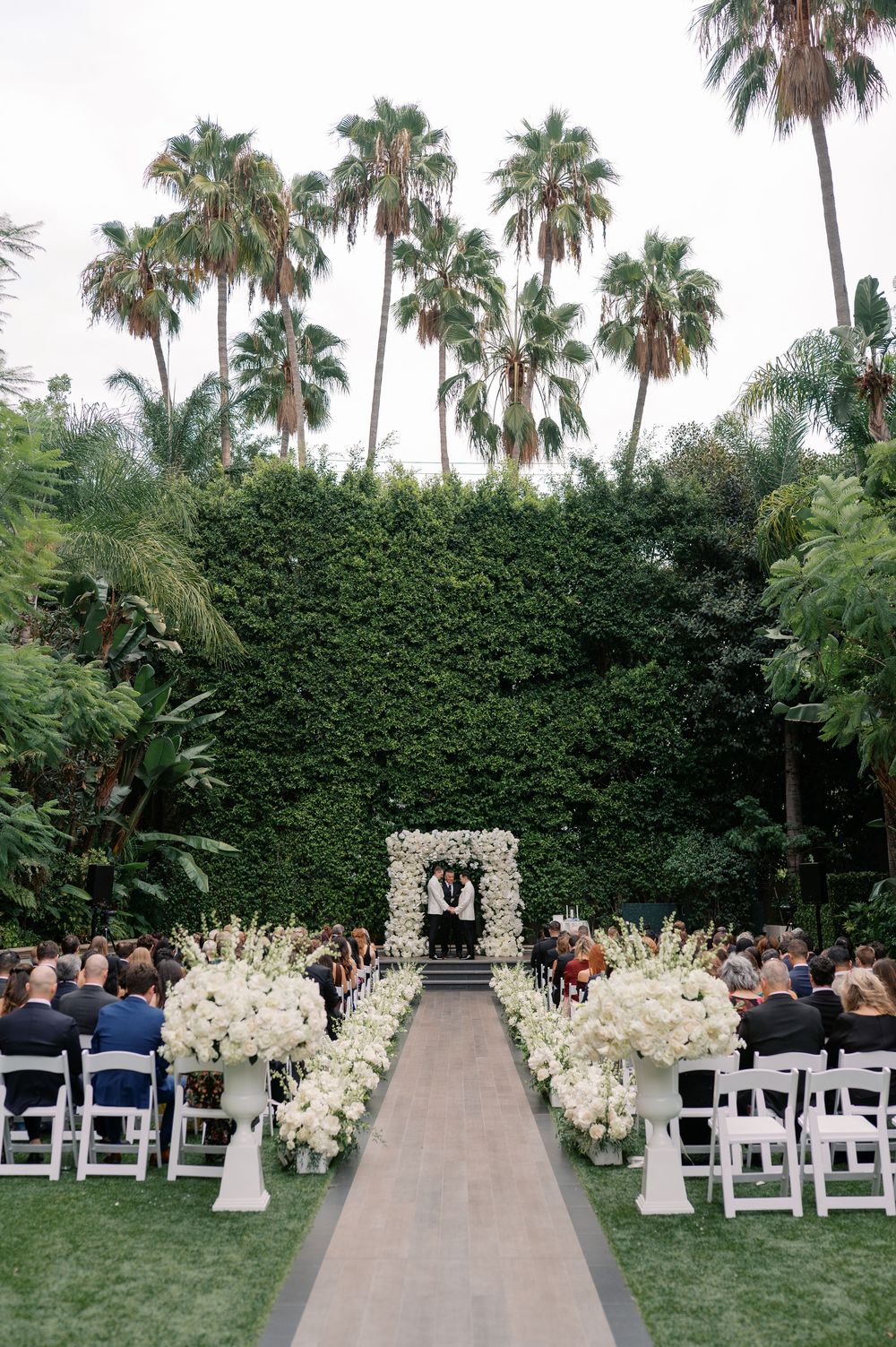 Elegant outdoor wedding ceremony with white floral arrangements lining the aisle, arch, and lush green hedge backdrop with tall palm trees.