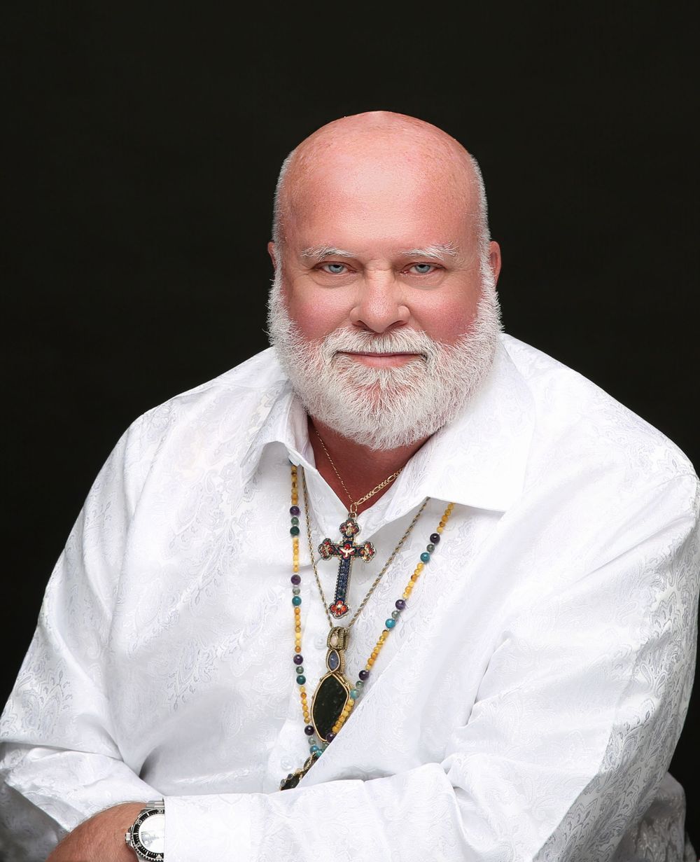 Man with a white beard in a white shirt and beaded necklaces on a black backdrop, classic portrait