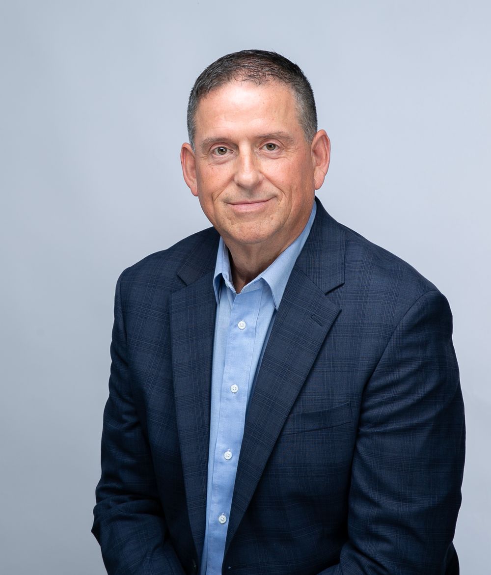 Man in a navy blazer and light blue shirt on a light gray backdrop, corporate headshot for men