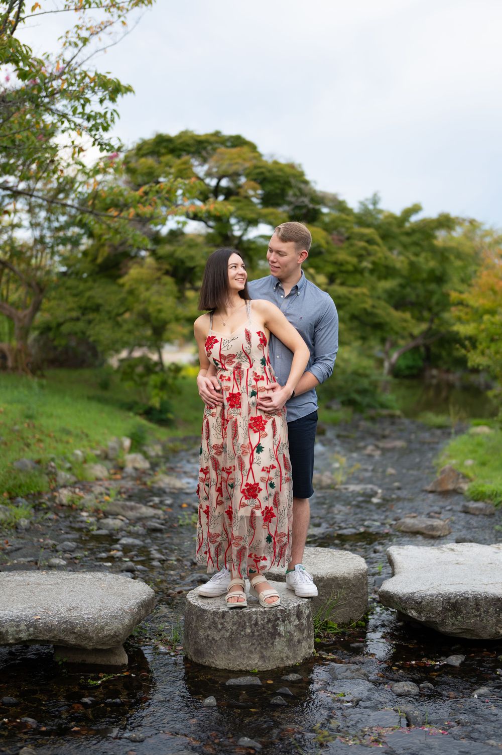 Pregnant couple standing on stepping stones over a stream, surrounded by lush green trees in a serene outdoor setting.