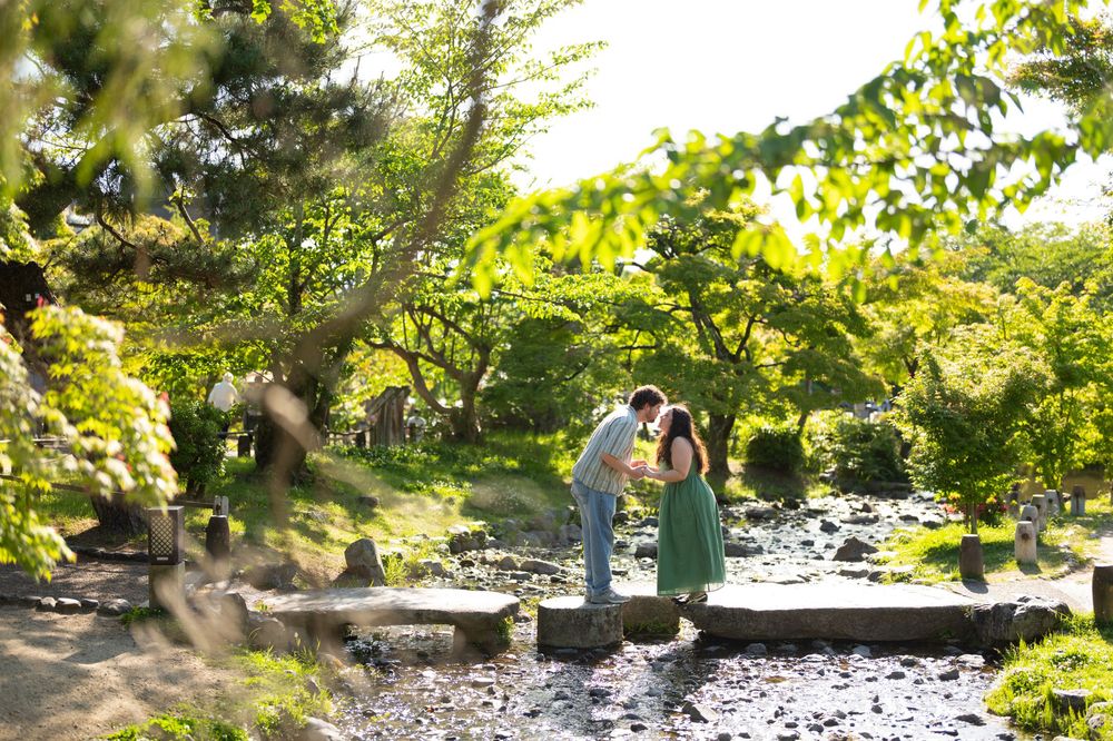 Couple sharing a romantic moment on stepping stones in a lush Japanese garden with green trees and a tranquil stream.