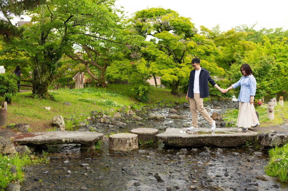 Couple holding hands crossing stepping stones over a stream in a lush Japanese garden.