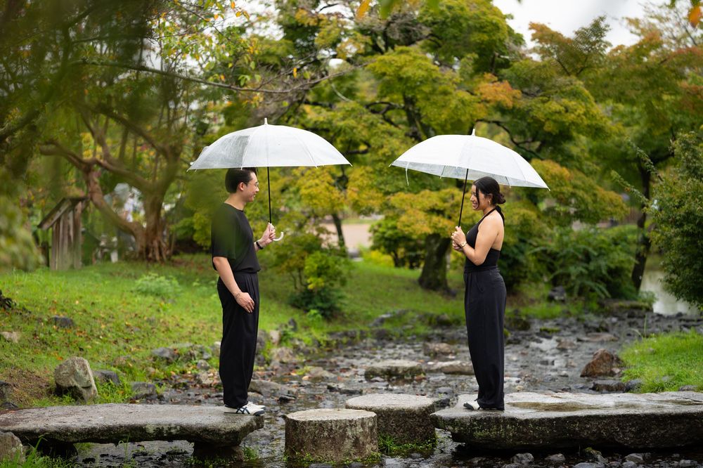 Couple holding white umbrellas standing on stepping stones in a lush green garden during a rainy day.