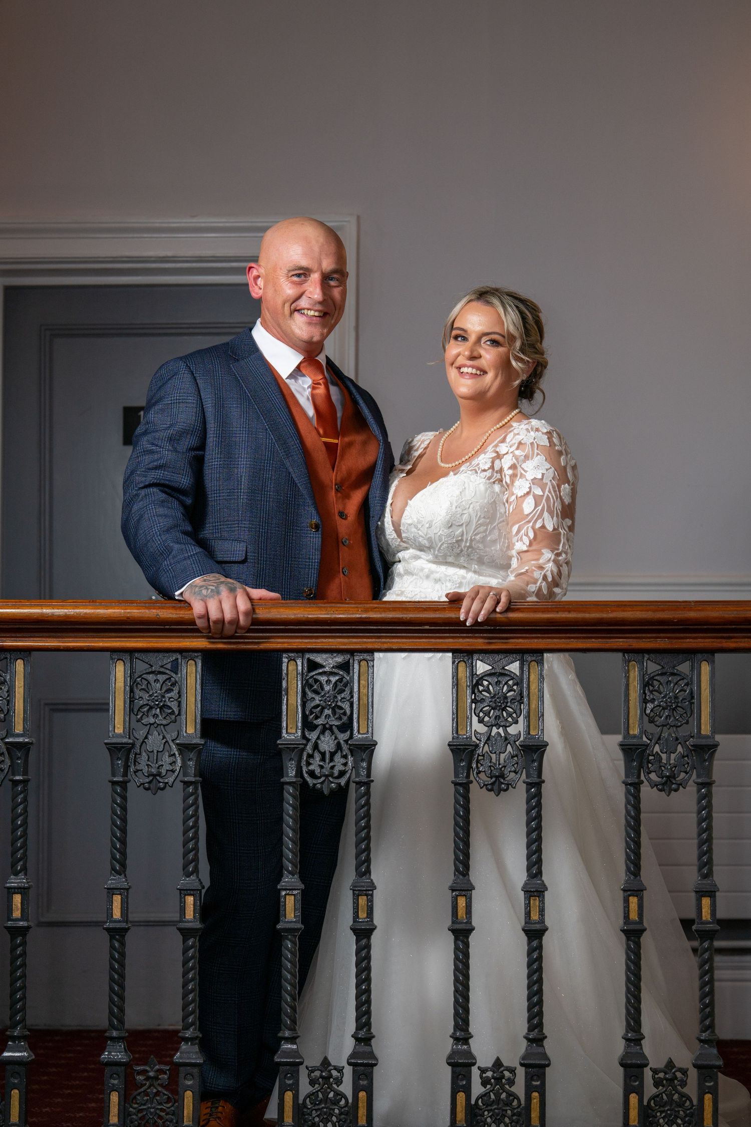 Smiling bride and groom pose elegantly at an ornate staircase railing, she in lace gown, he in navy suit with rust tie.