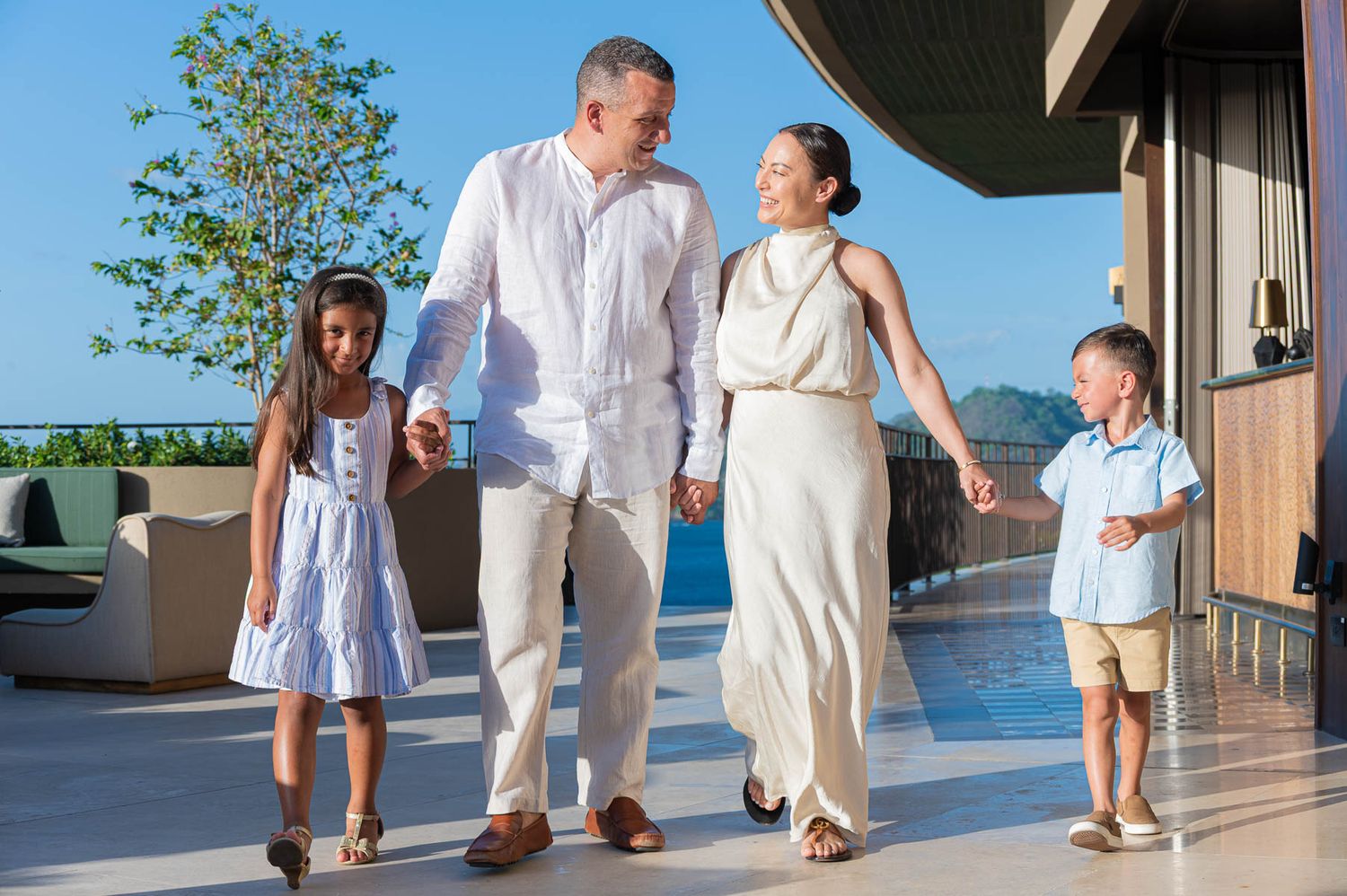 Smiling family of four in light summer outfits walking hand in hand along a sunny resort terrace with ocean and mountain views.