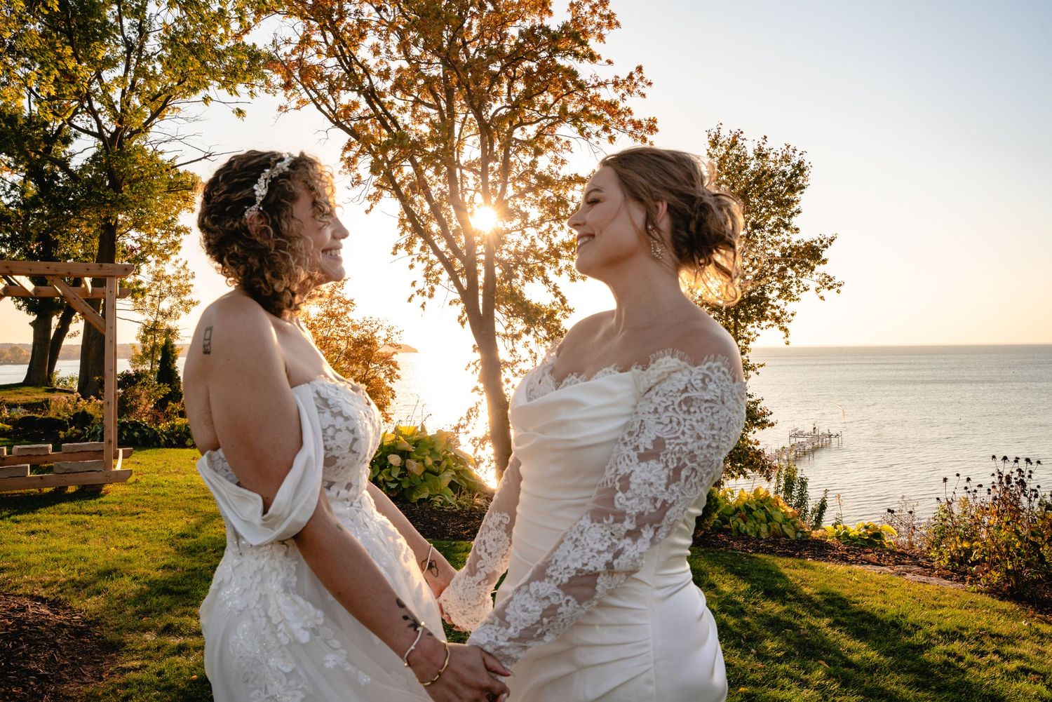 Two brides in white lace wedding gowns hold hands at golden hour by the waterfront.