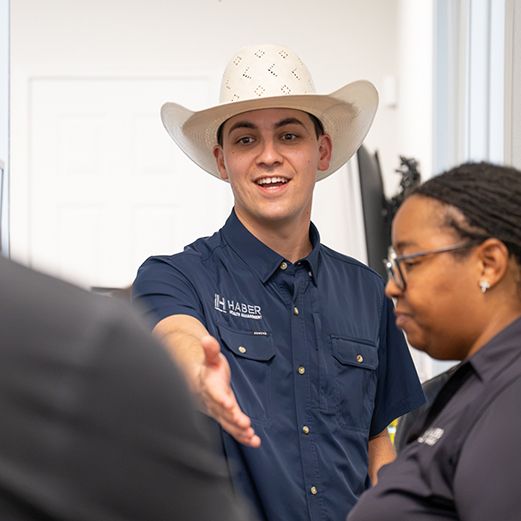 Smiling young man in a white straw cowboy hat and navy Habbir work shirt extends his hand to greet colleagues in a bright office.
