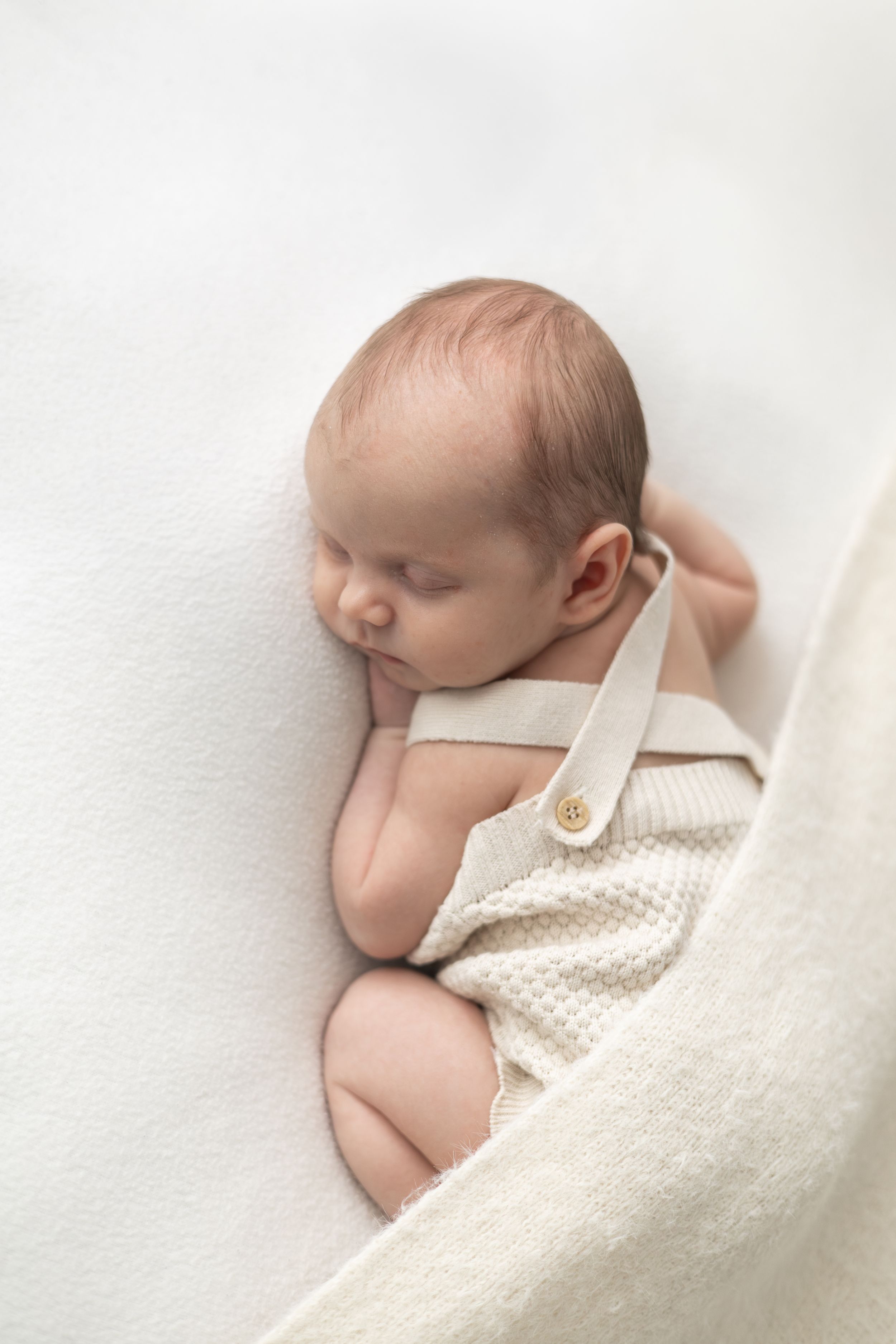 Peaceful newborn baby sleeping on a white blanket wearing a cream knit romper.
