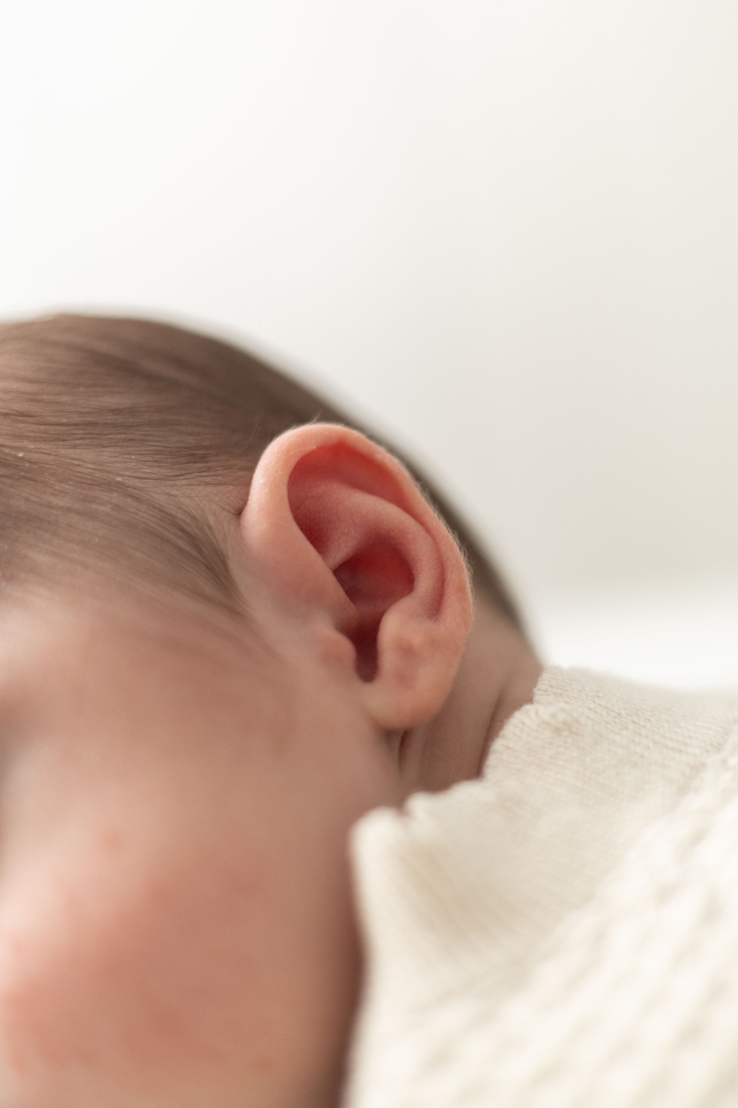 Close-up of a newborn baby's tiny ear with soft skin and fine hair, resting on a white blanket.