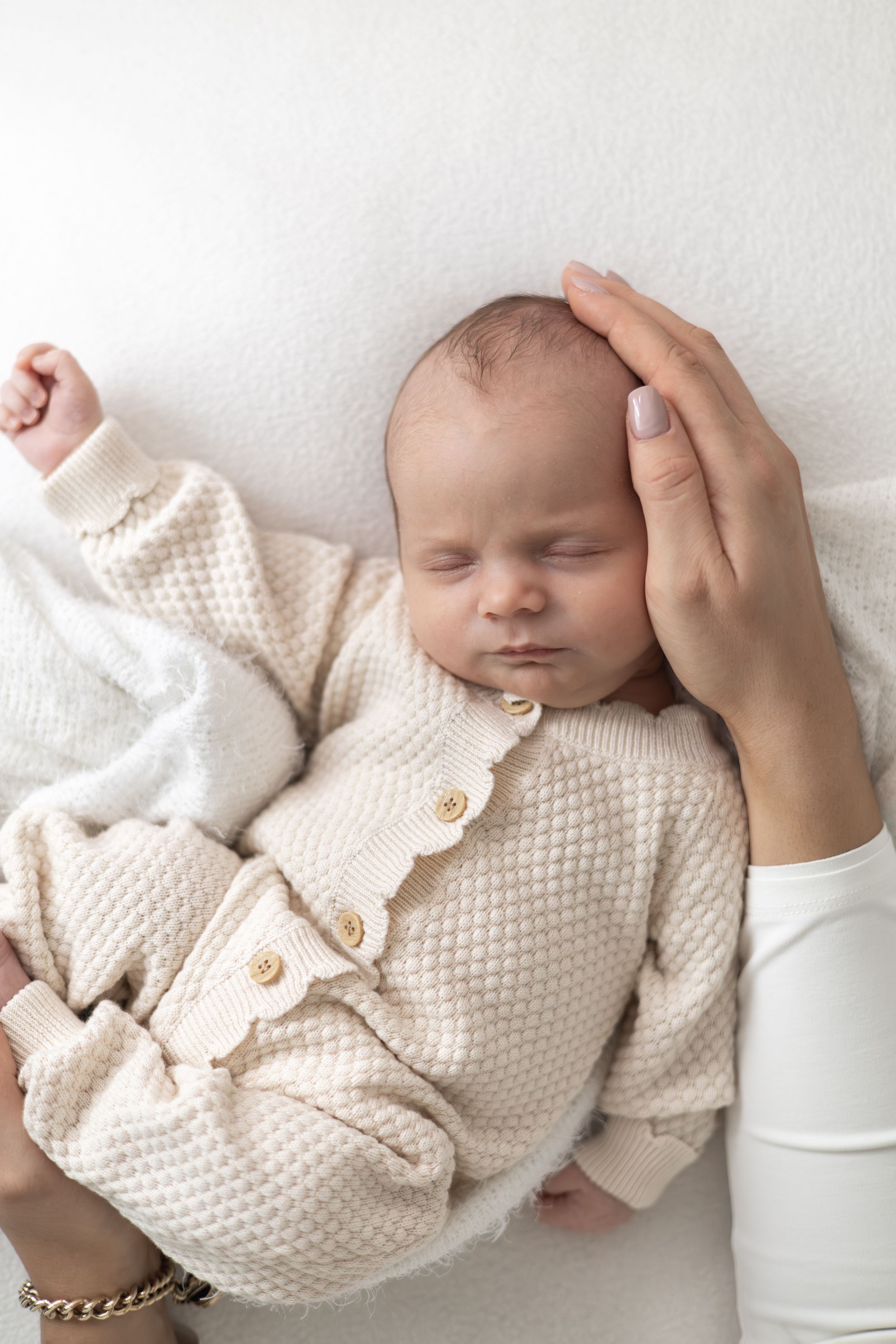 Newborn baby sleeping peacefully in a cream knit outfit while mums gentle hand cradles their head on a white background.