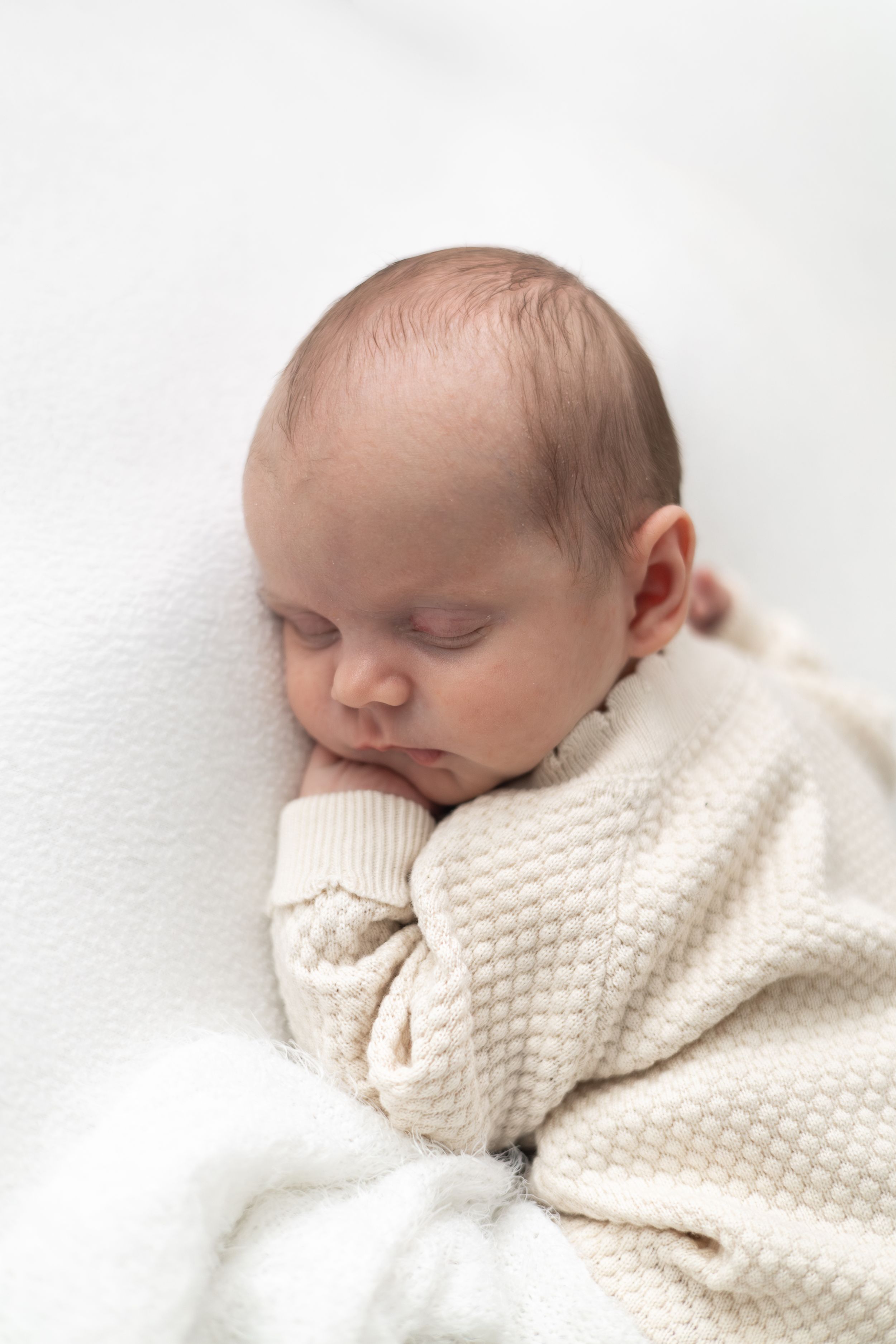 A peaceful newborn baby sleeping wrapped in a cream knit blanket on a soft white background.