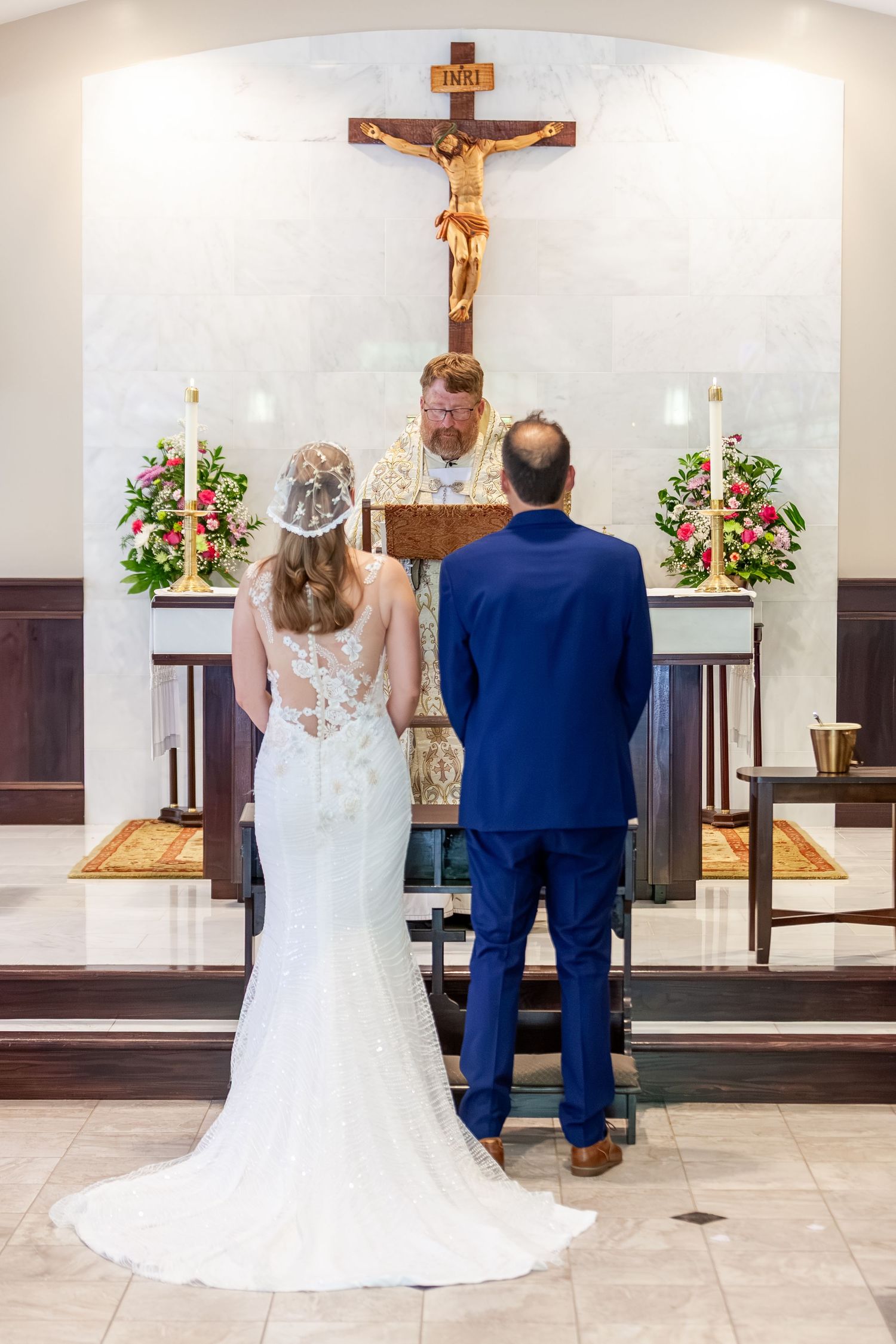 Bride and groom stand at church altar before priest during Catholic wedding ceremony with crucifix backdrop.