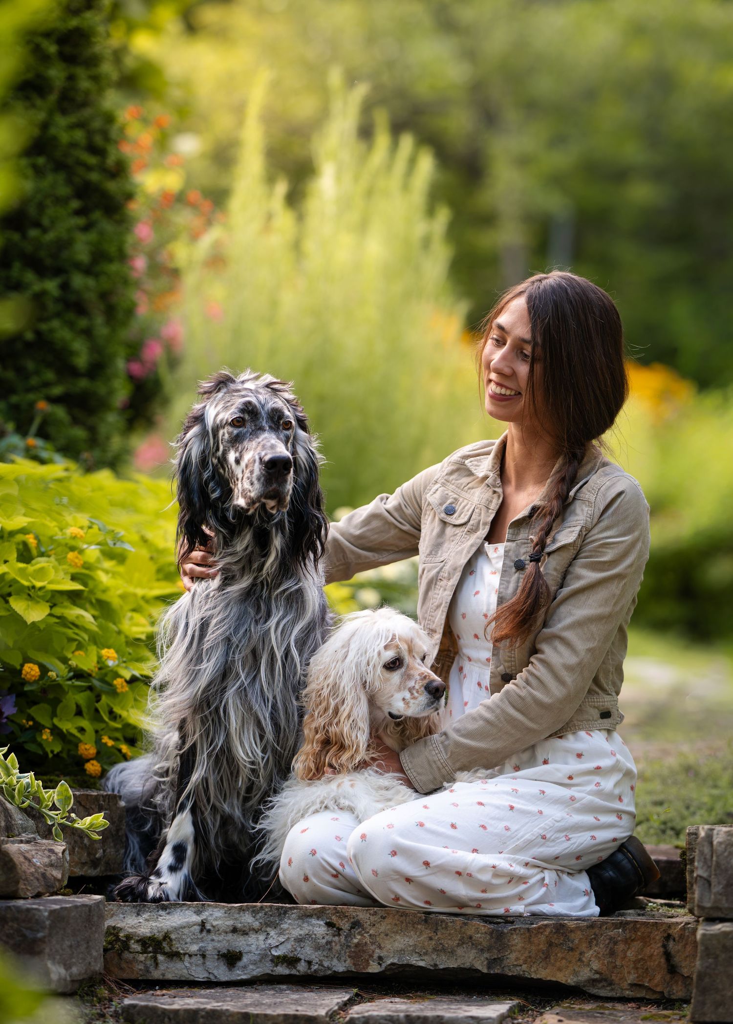 Femme souriante assise sur marches de pierre avec son setter anglais et son cocker, dans un jardin fleuri lors d'une séance photo animaliere