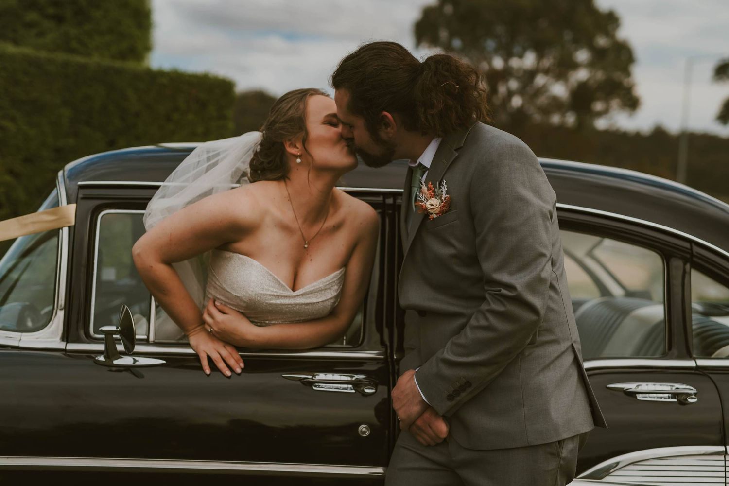 Bride and groom share a romantic kiss leaning against a classic black vintage car on their wedding day.