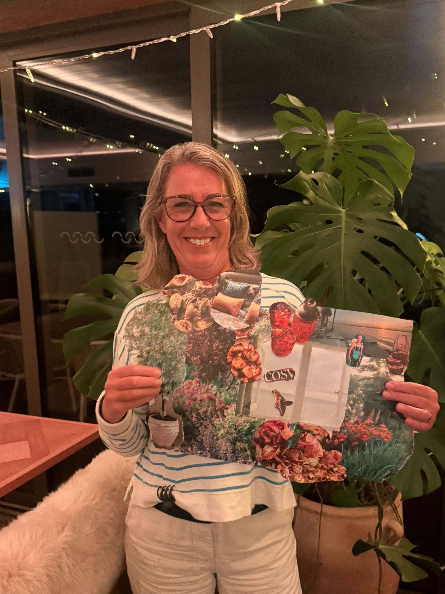 Woman smiling and holding a floral magazine inside a restaurant with tropical plants and string lights.